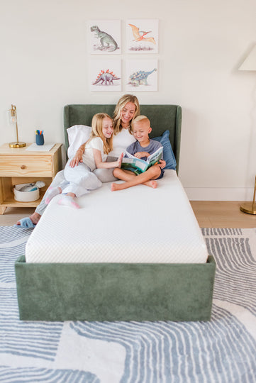 Family reading on a comfortable mattress in a cozy bedroom setting with dinosaur wall art.