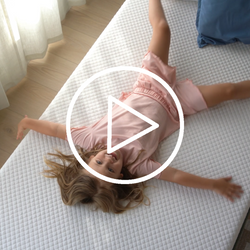 Child in pink pajamas playfully lying on a comfortable white mattress in a sunny bedroom.