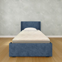 Blue upholstered single bed with white mattress and pillow on hardwood floor against a light textured wall