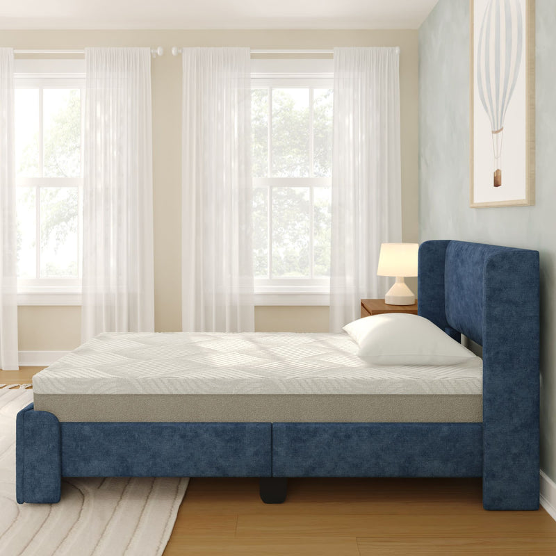 Blue upholstered platform bed with memory-foam mattress and white pillow in bright modern bedroom with sheer curtains, wooden nightstand, lamp and framed wall art