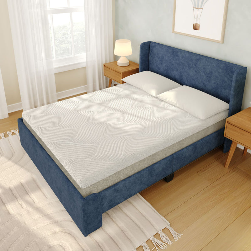 White textured mattress topper on blue upholstered bed frame with two pillows, wooden nightstands and lamp in bright modern bedroom with hardwood floor, area rug and sheer window curtains