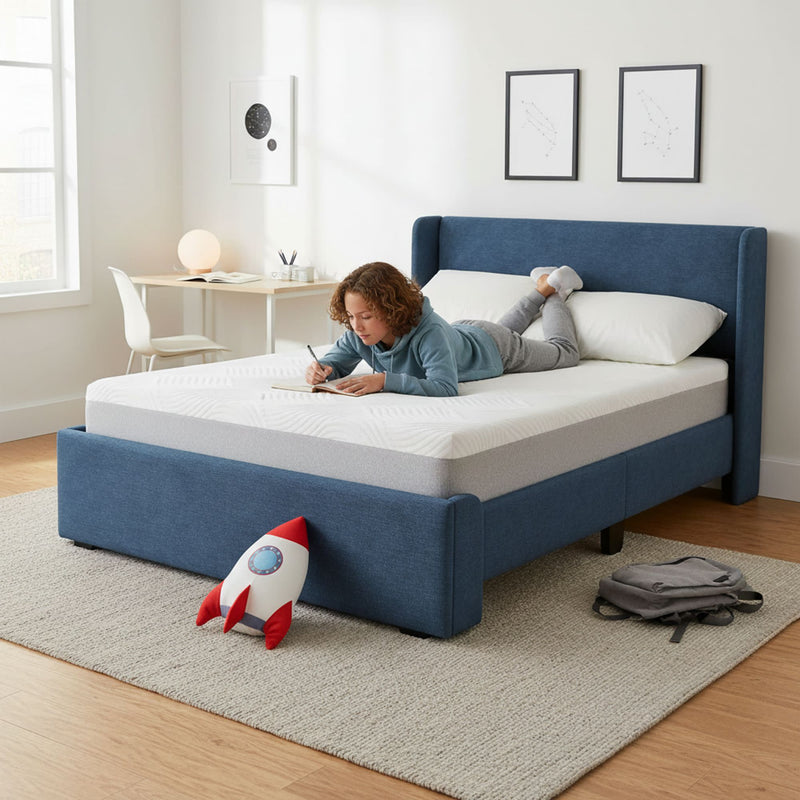 Modern kids bedroom with blue upholstered bed and white mattress, child writing in notebook on bed, study desk, rocket plush toy and backpack on rug