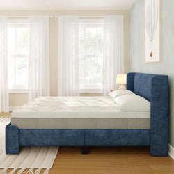 Sunlit modern bedroom featuring a blue upholstered bed frame with mattress and white pillows, bedside lamp, sheer white curtains, hardwood floor and area rug