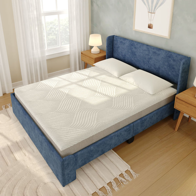 Blue upholstered platform bed with white memory foam mattress and pillows in a bright modern bedroom with wooden nightstands, hardwood floor, neutral rug and sheer curtains