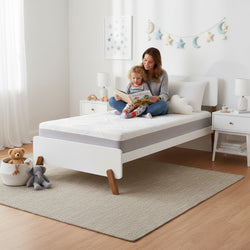 Mother reading to toddler on modern white twin bed with grey mattress in bright nursery featuring star-and-moon wall decor, plush toys, bedside tables and neutral rug
