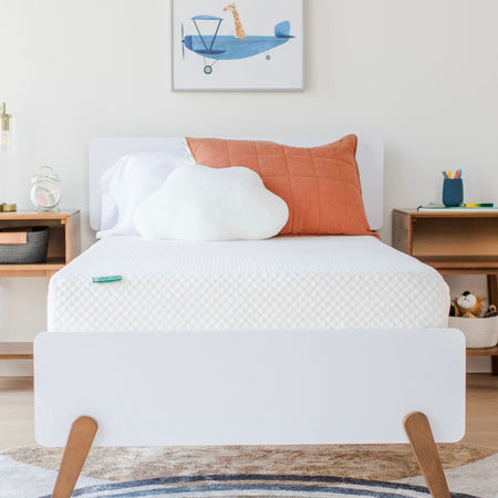 Modern kids bedroom with white bed, cloud-shaped pillow, and orange cushion on a comfortable mattress, featuring a playful airplane wall art.